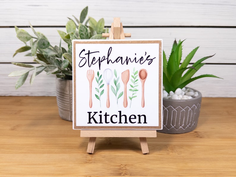 K&ouml;nnte beinhalten: Ein wei&szlig;es Schild mit dem Text "Stephanie's Kitchen" in schwarzen Buchstaben. Das Schild ist mit einer Aquarell-Illustration von K&uuml;chenutensilien und Gr&uuml;npflanzen verziert. Das Schild wird auf einem kleinen Holzst&auml;nder pr&auml;sentiert.