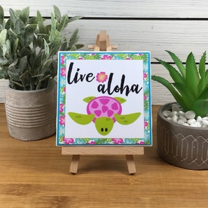 May include: A small wooden easel displaying a square sign with a tropical design. The sign features a pink and green sea turtle with the text "Live Aloha" in black lettering.