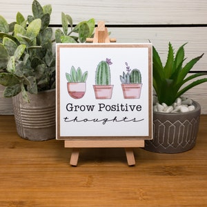 May include: A small wooden sign with a white background and a brown border. The sign has a hand-drawn illustration of three potted cacti with pink flowers. The text "Grow Positive thoughts" is written in black cursive below the cacti.