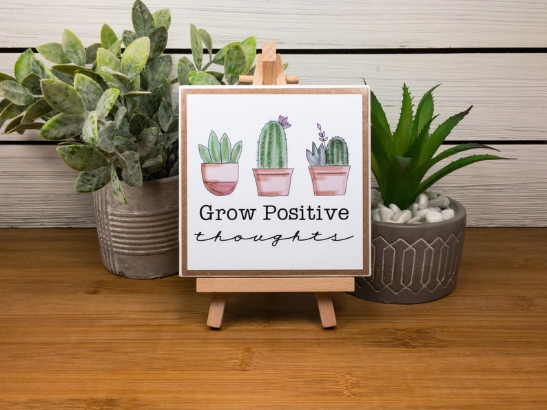 May include: A small wooden sign with a white background and a brown border. The sign has a hand-drawn illustration of three potted cacti with pink flowers. The text "Grow Positive thoughts" is written in black cursive below the cacti.