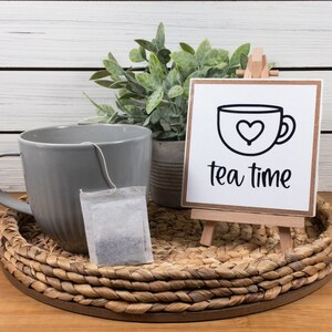 May include: A gray ceramic mug with a tea bag sits on a woven tray next to a small wooden easel with a white sign that reads "tea time" in black lettering. A potted green plant is behind the tray.