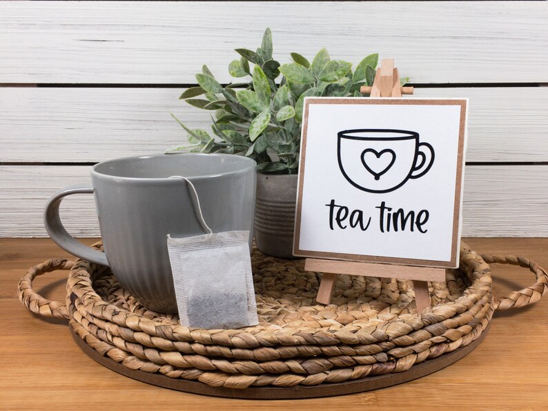 May include: A gray ceramic mug with a tea bag sits on a woven tray next to a small wooden easel with a white sign that reads "tea time" in black lettering. A potted green plant is behind the tray.