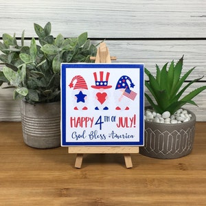 May include: A small wooden sign with a blue background and three gnomes wearing red, white, and blue hats. The sign says "Happy 4th of July! God Bless America".