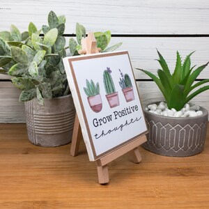 May include: A white ceramic tile with a brown wooden easel stand. The tile has a hand-drawn illustration of three potted succulents with the text "Grow Positive Thoughts".
