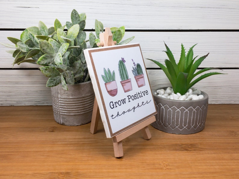 May include: A white ceramic tile with a brown wooden easel stand. The tile has a hand-drawn illustration of three potted succulents with the text "Grow Positive Thoughts".