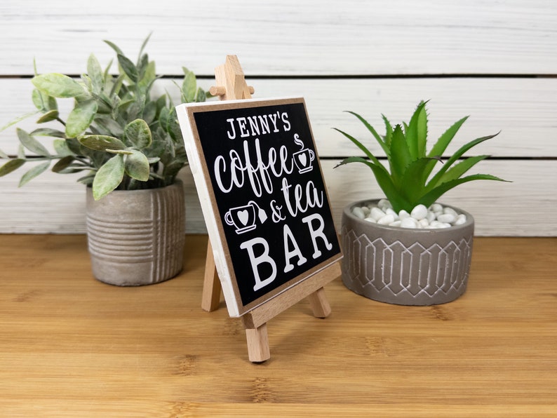 May include: A small black sign with white text on a wooden easel. The sign reads "Jenny's Coffee & Tea Bar".
