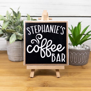 May include: A small black chalkboard sign with white text that reads "Stephanie's Coffee Bar" on a wooden easel. The sign is sitting on a wooden table with two potted plants.