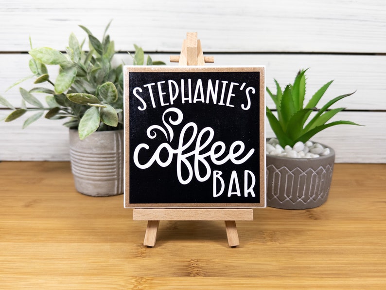 May include: A small black chalkboard sign with white text that reads "Stephanie's Coffee Bar" on a wooden easel. The sign is sitting on a wooden table with two potted plants.