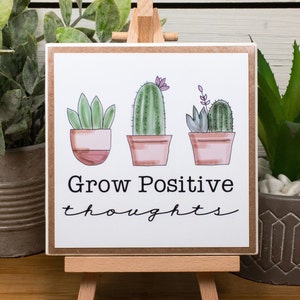 May include: A white sign with a brown border featuring three potted cacti with the text "Grow Positive thoughts" in black.