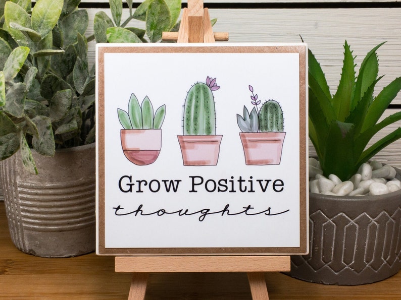 May include: A white sign with a brown border featuring three potted cacti with the text "Grow Positive thoughts" in black.