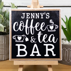 May include: A black wooden sign with white text that reads "Jenny's coffee & tea bar" with a small coffee cup and teacup graphic. The sign is on a small wooden easel.