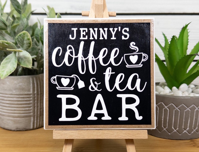 May include: A black wooden sign with white text that reads "Jenny's coffee & tea bar" with a small coffee cup and teacup graphic. The sign is on a small wooden easel.