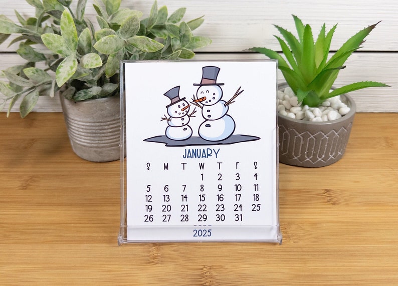 May include: A white desk calendar with a January 2025 calendar grid. The calendar features a cartoon illustration of two snowmen with black top hats and carrot noses. The snowmen are standing on a blue background.