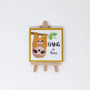 Puede incluir: Una impresi&oacute;n cuadrada con un borde amarillo, que representa dos perezosos de dibujos animados colgando de una rama, con las palabras "HANG in there". La impresi&oacute;n se muestra en un peque&ntilde;o caballete de madera.
