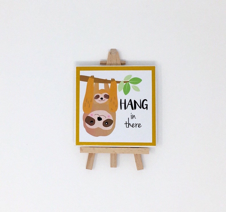 Puede incluir: Una impresi&oacute;n cuadrada con un borde amarillo, que representa dos perezosos de dibujos animados colgando de una rama, con las palabras "HANG in there". La impresi&oacute;n se muestra en un peque&ntilde;o caballete de madera.