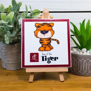 May include: A small, white square tile with a red border features a cartoon tiger with orange and black stripes. The tile is on a wooden easel and says "Year of the Tiger" in black text. The Chinese character for tiger is in red on the tile.