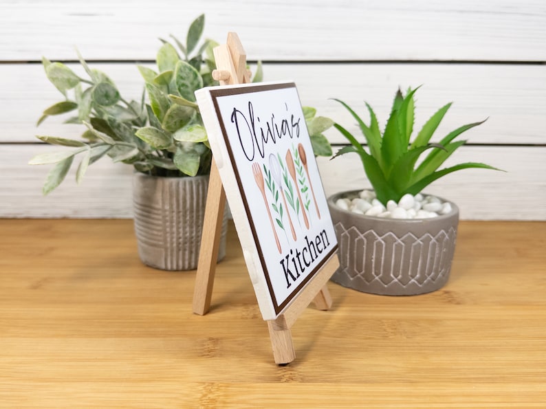 K&ouml;nnte beinhalten: Ein wei&szlig;es quadratisches Schild mit dem Text "Olivia's Kitchen" und einer Aquarell-Illustration von K&uuml;chenutensilien und Gr&uuml;npflanzen. Das Schild wird auf einem kleinen Holzstaffelei pr&auml;sentiert.