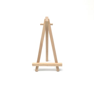 Puede incluir: Un peque&ntilde;o caballete de madera natural, con un dise&ntilde;o de tres patas. El caballete est&aacute; hecho de madera de color claro y presenta una barra horizontal en la base para sostener un lienzo o una obra de arte. El caballete est&aacute; dise&ntilde;ado para colocarse sobre una superficie plana.