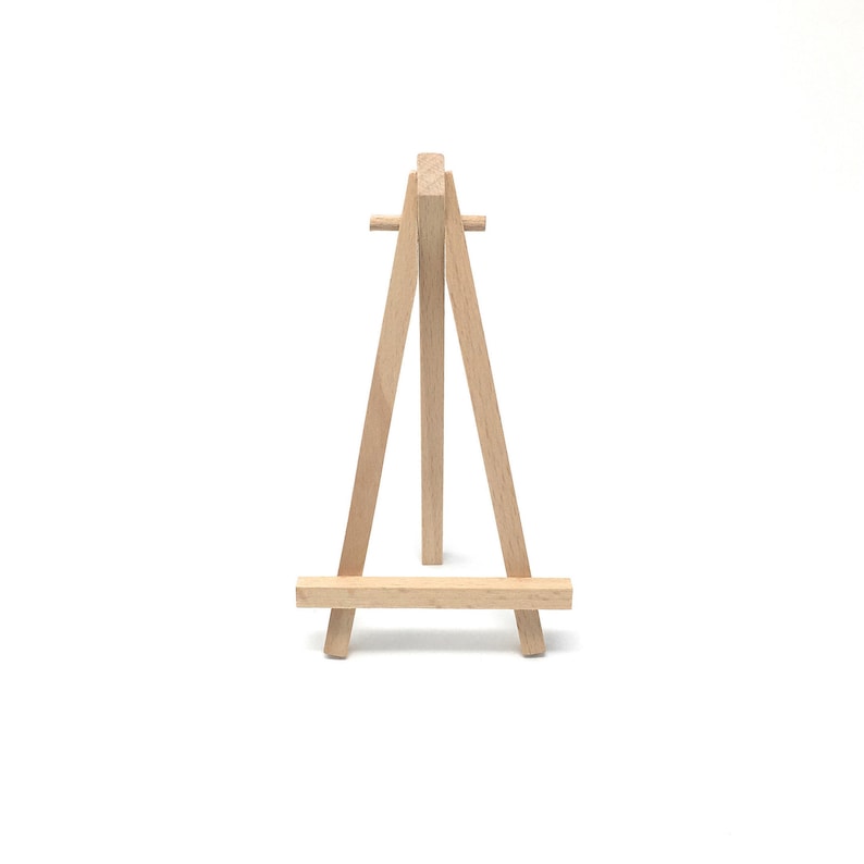 Puede incluir: Un peque&ntilde;o caballete de madera natural, con un dise&ntilde;o de tres patas. El caballete est&aacute; hecho de madera de color claro y presenta una barra horizontal en la base para sostener un lienzo o una obra de arte. El caballete est&aacute; dise&ntilde;ado para colocarse sobre una superficie plana.