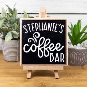 May include: A small black chalkboard sign with white text that reads "Stephanie's Coffee Bar" on a wooden easel.
