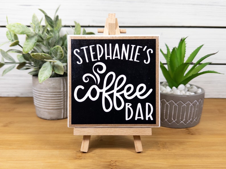 May include: A small black chalkboard sign with white text that reads "Stephanie's Coffee Bar" on a wooden easel.