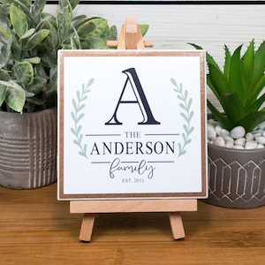 May include: A white square sign with a brown wood frame on a small wooden easel. The sign has a green leafy design with the letter A in black and the text "THE ANDERSON family EST. 2011".