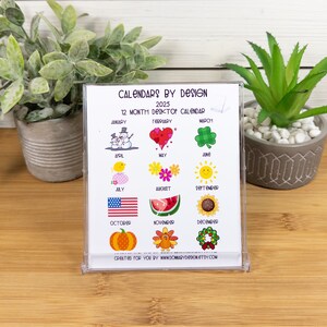 May include: A white desktop calendar with a clear plastic frame. The calendar features a different colorful illustration for each month of the year. The text "CALENDARS BY DESIGN" and "2025" are printed at the top of the calendar. The text "12 MONTH DESKTOP CALENDAR" is printed below the title. The text "CREATED FOR YOU BY WWW.DOMARYDDONETTY.COM" is printed at the bottom of the calendar.