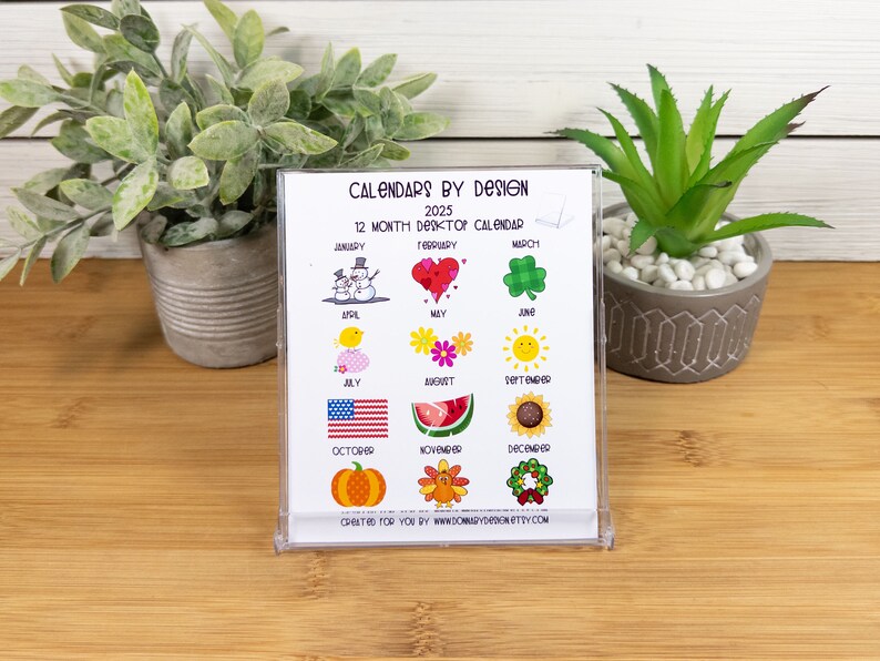 May include: A white desktop calendar with a clear plastic frame. The calendar features a different colorful illustration for each month of the year. The text "CALENDARS BY DESIGN" and "2025" are printed at the top of the calendar. The text "12 MONTH DESKTOP CALENDAR" is printed below the title. The text "CREATED FOR YOU BY WWW.DOMARYDDONETTY.COM" is printed at the bottom of the calendar.