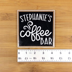 May include: A black square sign with white text that reads "Stephanie's coffee bar" with a stylized coffee steam design. The sign has a light brown wood frame.