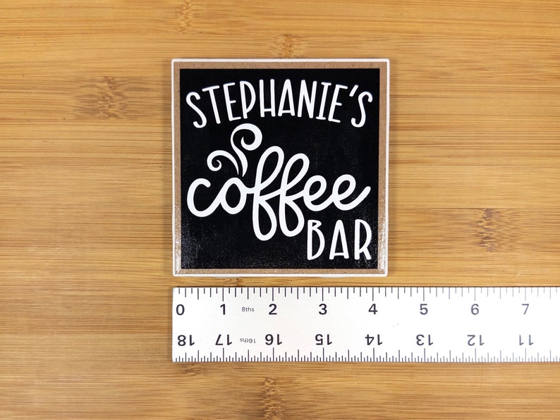 May include: A black square sign with white text that reads "Stephanie's coffee bar" with a stylized coffee steam design. The sign has a light brown wood frame.