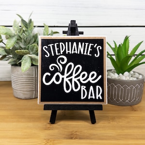 May include: Black and brown wooden sign with white text that reads "Stephanie's Coffee Bar" with a stylized coffee steam design. The sign is on a small black easel.