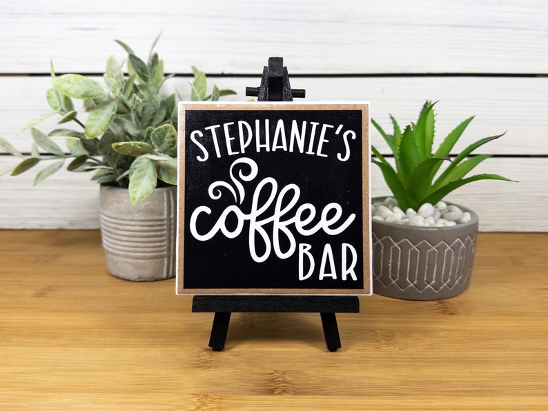 May include: Black and brown wooden sign with white text that reads "Stephanie's Coffee Bar" with a stylized coffee steam design. The sign is on a small black easel.