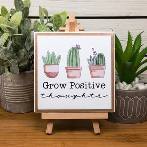 May include: A wooden sign with a white background and a brown border. The sign features three potted cacti with pink flowers and the text "Grow Positive thoughts". The sign is displayed on a small wooden easel.