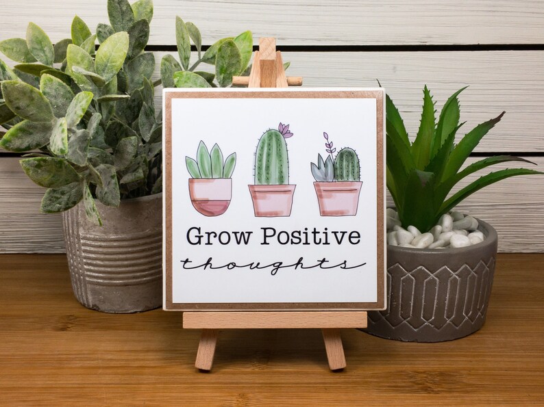May include: A wooden sign with a white background and a brown border. The sign features three potted cacti with pink flowers and the text "Grow Positive thoughts". The sign is displayed on a small wooden easel.