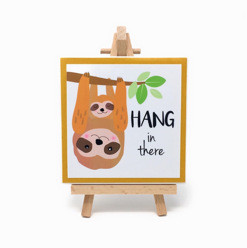 Puede incluir: Una impresi&oacute;n cuadrada enmarcada en un peque&ntilde;o caballete de madera. La impresi&oacute;n presenta dos perezosos de dibujos animados colgando de una rama, con las palabras "HANG in there" en texto negro. El marco es amarillo y los perezosos son marrones y tostados.
