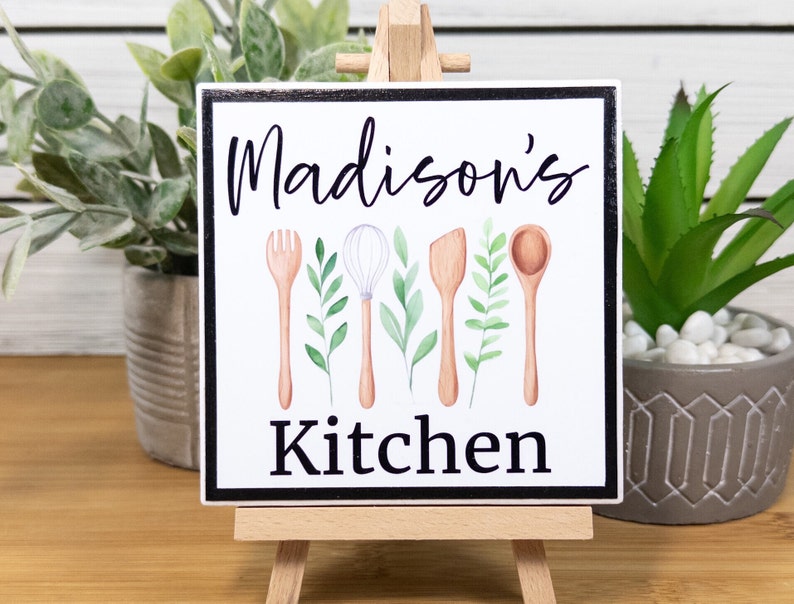 K&ouml;nnte beinhalten: Ein wei&szlig;es quadratisches Schild mit schwarzem Rahmen, auf dem in schwarzer Schrift "Madison's Kitchen" steht. Das Schild zeigt eine Aquarell-Illustration von K&uuml;chenutensilien, darunter eine Gabel, ein Schneebesen, ein Spatel und ein L&ouml;ffel, mit gr&uuml;nen Bl&auml;ttern.