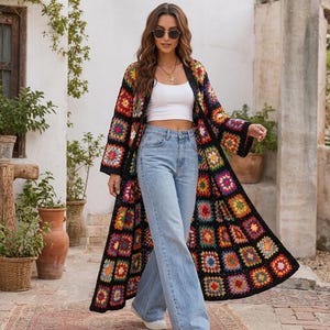 May include: A long, open-front cardigan made from a black-based granny square crochet pattern featuring vibrant, multi-colored floral motifs in shades of red, yellow, blue, and pink, worn over a white crop top and light blue denim jeans.