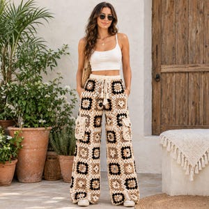 Pode incluir: Calças de cintura subida e perna larga com um padrão de crochet granny square em tons de creme, bege e preto. As calças incluem uma cintura com cordão e borlas, e dois bolsos laterais tipo cargo com fecho de botão, feitas de fio de malha.