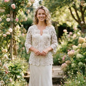 May include: A cream-colored, long-sleeved crochet cardigan featuring intricate floral patterns and scalloped edges, worn over a matching cream-colored slip dress that falls to the mid-calf, creating a coordinated ensemble with a textured finish.
