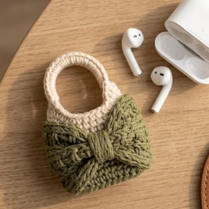 May include: A crochet protective case for wireless earbuds featuring a cream-coloured top section and a textured olive green bottom section adorned with a prominent central bow. Two white wireless earbuds and their open charging case are nearby.
