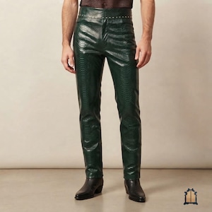 Puede incluir: Pantalones de color verde oscuro con textura de piel de serpiente y talle alto. La cintura está detallada con una hilera de pequeños remaches metálicos plateados. Los pantalones tienen un corte recto y ajustado, combinados con botas negras.