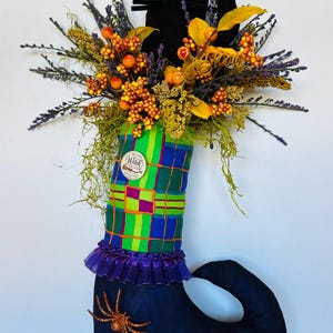 May include: A fabric witch boot decoration featuring a black base with a copper-colored spider, a purple ruffled trim, and a green and blue plaid upper section. It is topped with a black cat cutout and an arrangement of orange berries and lavender.
