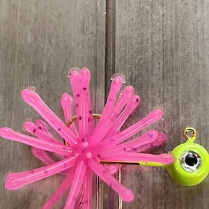 May include: A fishing lure featuring a bright neon pink soft plastic skirt with purple glitter flecks, attached to a chartreuse jig head with a single gold-colored hook and a prominent silver and black eye design on the side of the jig head.