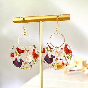 May include: A pair of gold hoop earrings with clear, semi-circular resin charms featuring a colorful chicken pattern. The earrings are displayed on a gold stand.