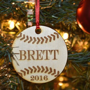 Op de afbeelding: Ronde houten kerstbal met een honkbalontwerp. De naam "BRETT" en het jaar "2016" zijn gegraveerd. Hangt aan een rood lint, perfect voor honkbalfans en feestelijke decoratie.