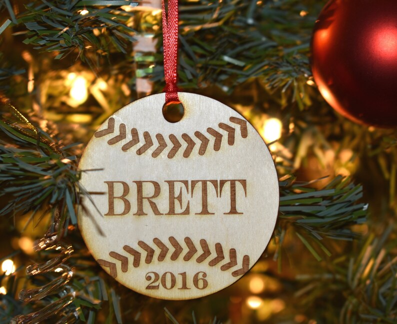 Op de afbeelding: Ronde houten kerstbal met een honkbalontwerp. De naam "BRETT" en het jaar "2016" zijn gegraveerd. Hangt aan een rood lint, perfect voor honkbalfans en feestelijke decoratie.