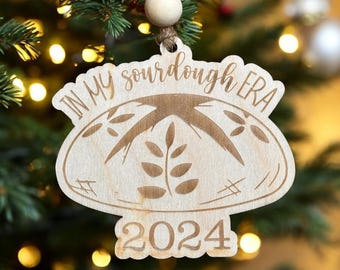 May include: A wooden Christmas ornament shaped like a loaf of bread with the text "In My Sourdough Era 2024" on it.