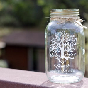 May include: A clear glass mason jar with a silver lid and twine tied around the neck. The jar features a white tree design with the names "Candice & Robert" and "French September 20, 2014" in a script font.