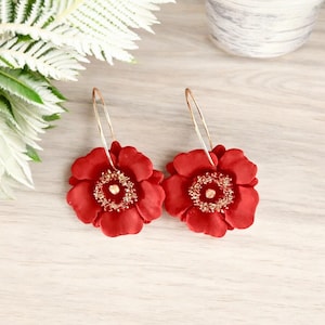 May include: A pair of hoop earrings featuring large, deep red flower-shaped charms. Each flower has layered petals and a textured, metallic gold-colored center. The thin, circular gold-toned wire hoops attach to the top of each floral piece.