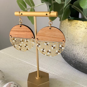 May include: A pair of gold tone earrings with a wood and clear resin design. The resin features a honeycomb pattern with small black and white bees.